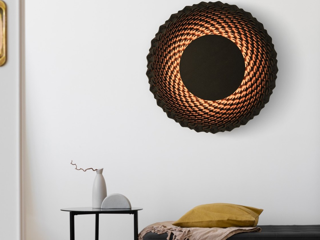 Decorative woven wall lamp with warm glow above a small round black table and a white vase in a minimalist bedroom corner.