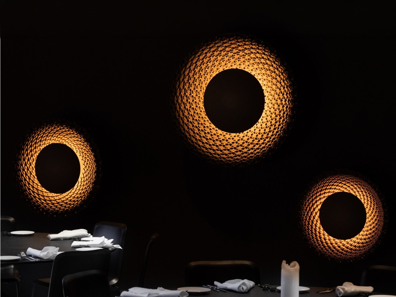 Dim dining room with three large circular woven wall lamps casting warm light over a table set with plates and napkins.