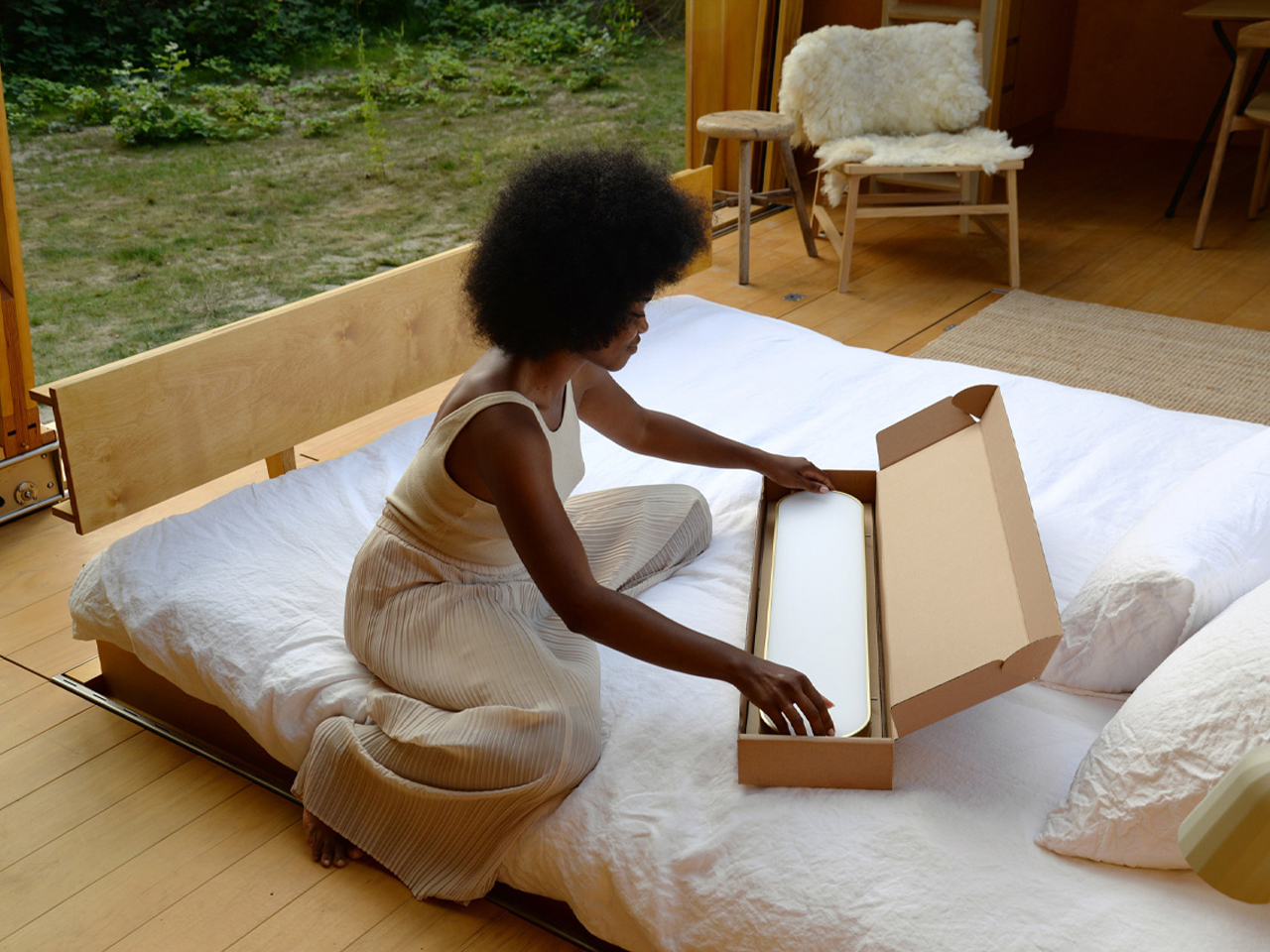 Woman with an afro sits on a bed and unboxes a long white item from a cardboard box in a bright wooden room.
