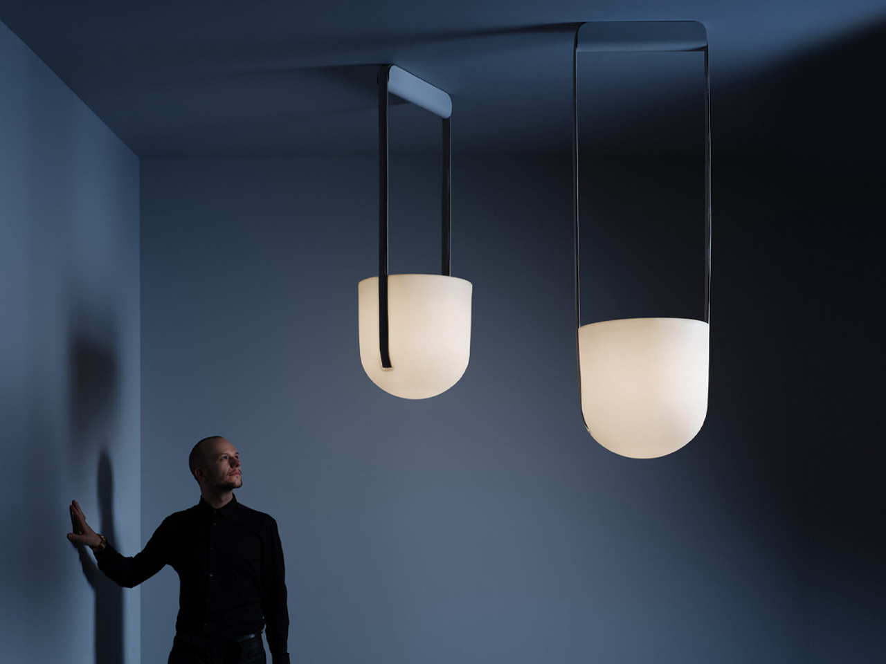 Man in black stands beside a blue wall, looking up at two modern frosted-glass pendant lights suspended from the ceiling.