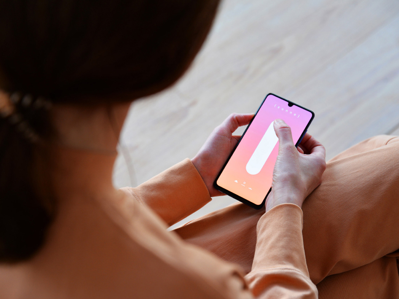 Person wearing peach clothing holds a smartphone with a pink gradient wallpaper and a white vertical oval shape on screen.