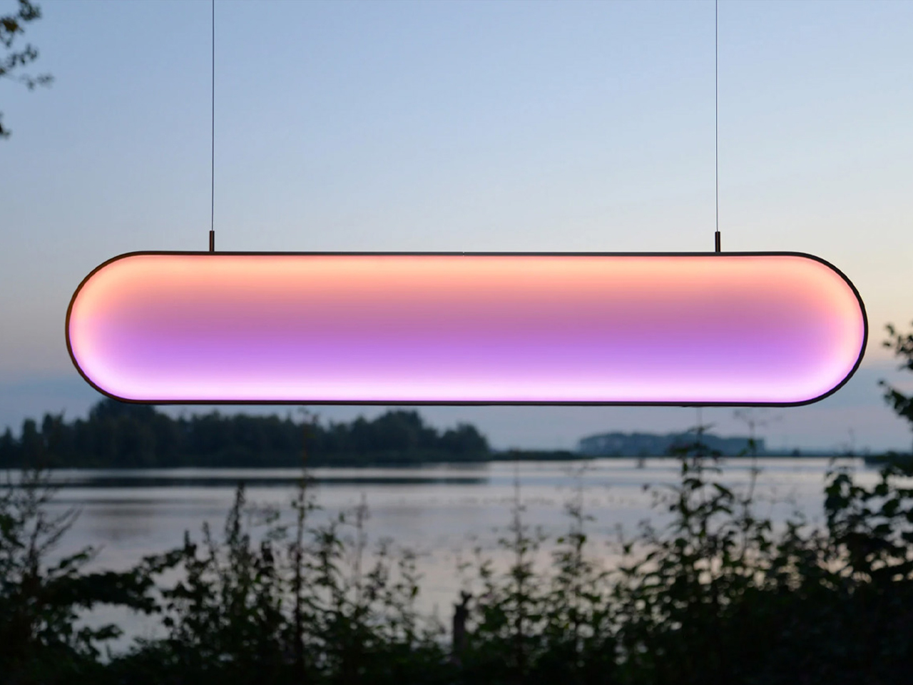 Hanging oval LED light fixture with pink-to-purple gradient, suspended by two cables over a lakeside scene at dusk.