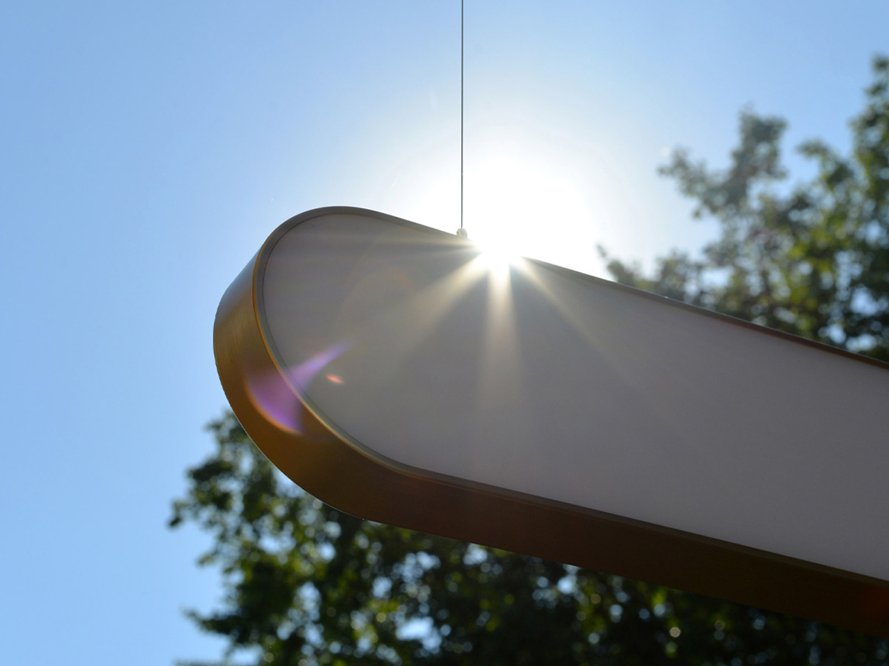 Sunlight streams over a white curved outdoor surface (likely sculpture or structure) with a bright flare against a clear blue sky and trees in the background, suggesting an outdoor installation or playground element.