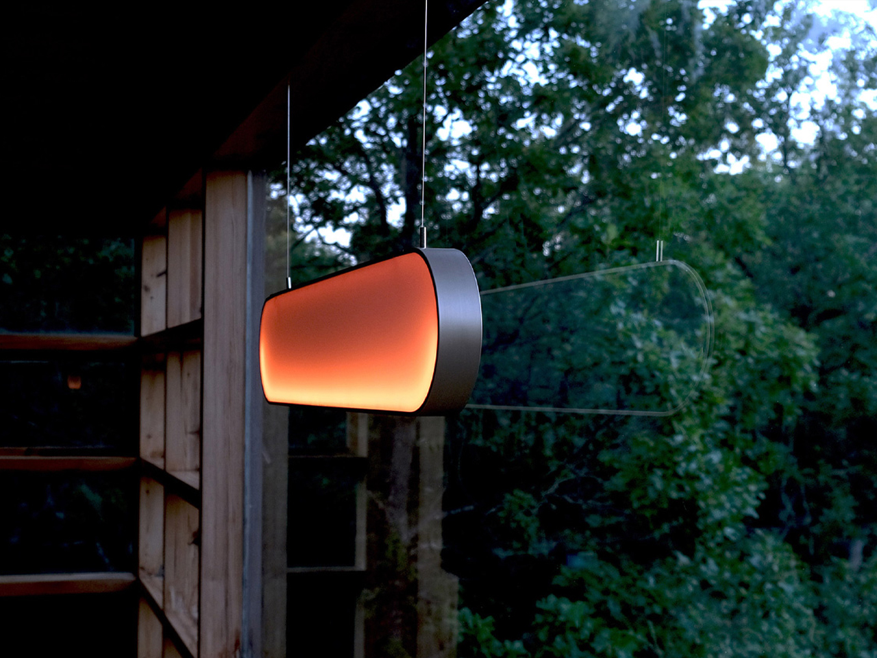 Outdoor hanging light fixture with a warm amber glow, suspended in front of a wooden structure and green foliage at dusk/evening.