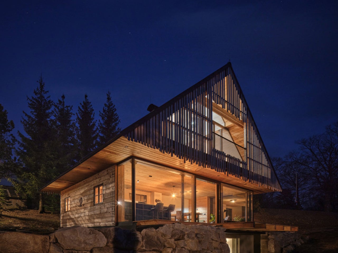 Modern timber house with a steep triangular roof, glass walls, and warm interior lights at night.