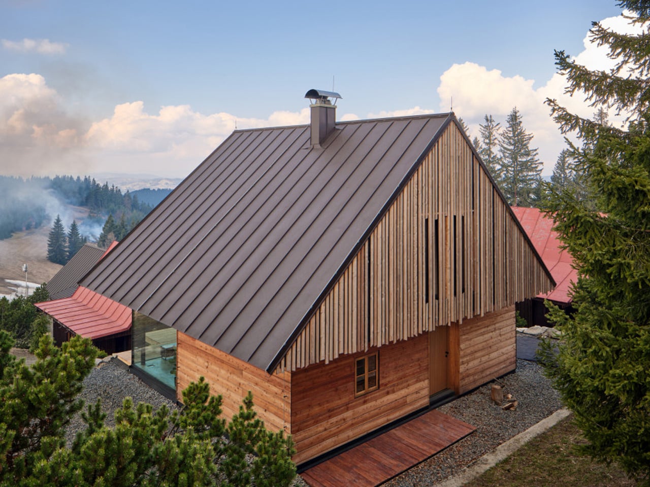 Wooden cabin with a steep metal roof in a forested mountain landscape.