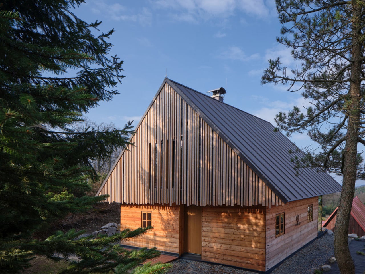 Wooden cabin with a steep metal roof, framed by pine trees under a blue sky