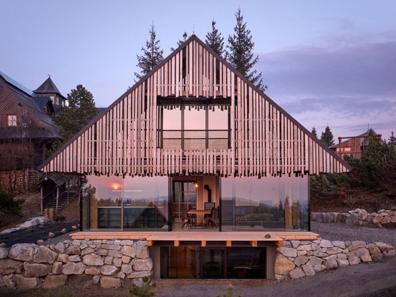 A modern A-frame cabin with vertical wooden slats on the upper story, glass walls on the ground floor, and a stone retaining wall surrounding the base at sunset.