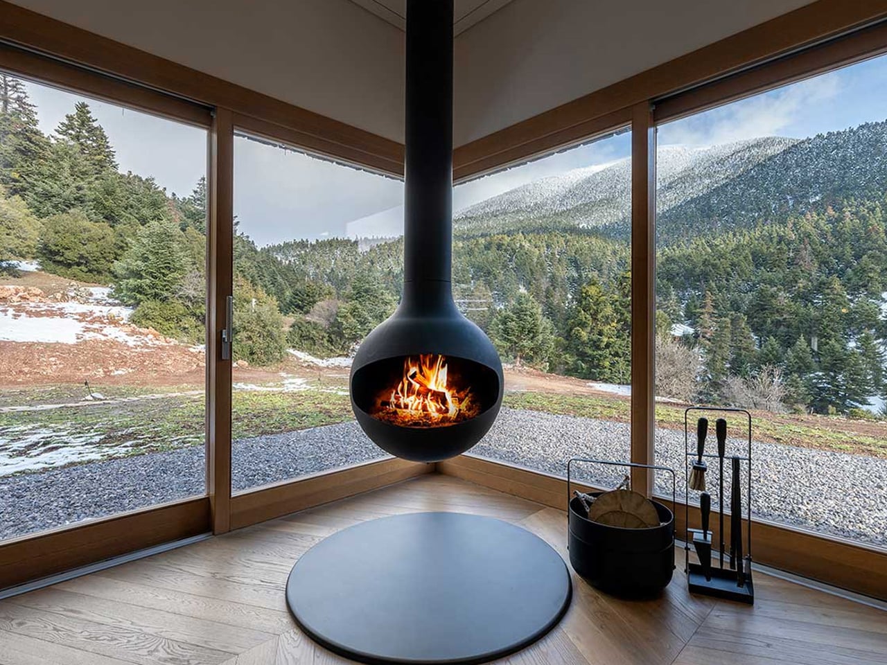 Suspended black spherical fireplace hanging in a corner with panoramic forest and snow outside, flame visible inside the stove.