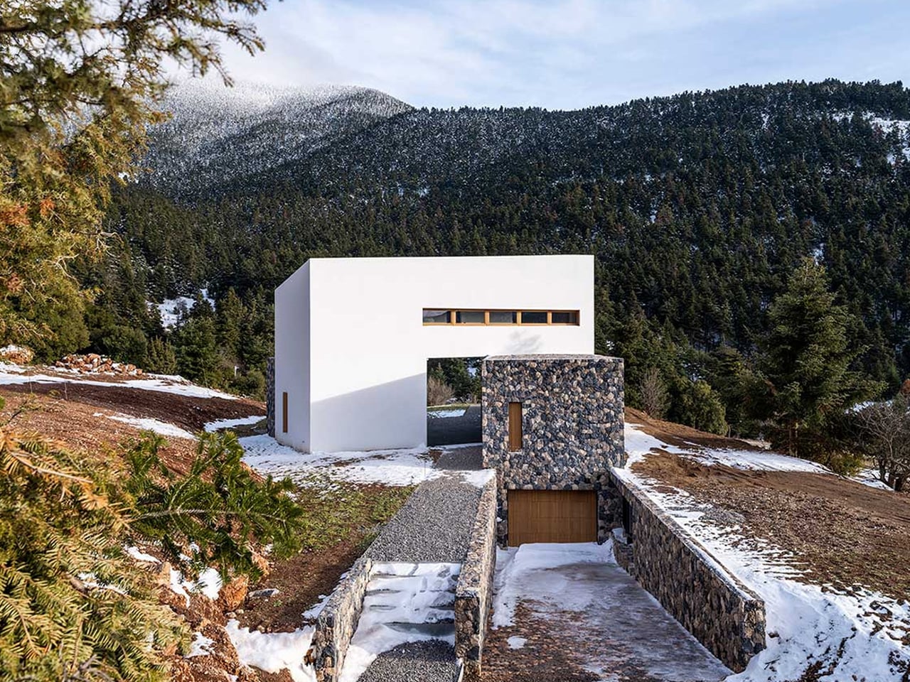Modern white cubic house with a stone tower and driveway leading to a wooden garage, set in a snowy hillside forest. (Informative)