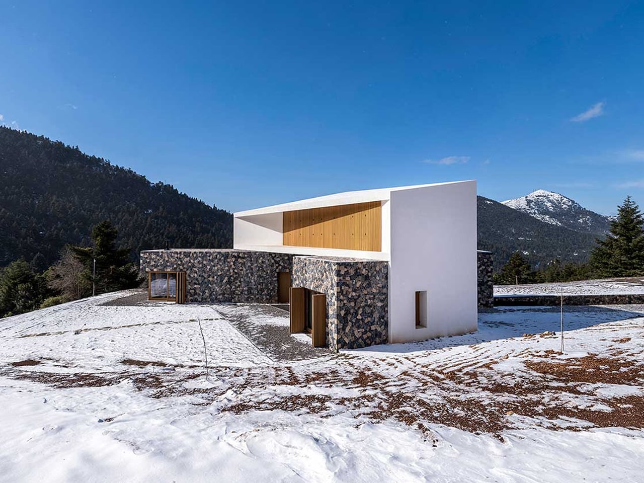 Contemporary two-story house with white and stone exterior sits on a snowy hillside against a mountainous backdrop.