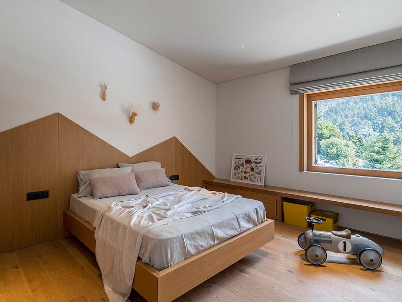 Modern bedroom with a low wooden bed, angled headboard, light bedding, and decorative deer silhouettes on the wall.