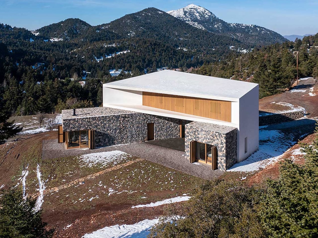 Modern house with white angular upper block and stone lower walls on a snowy hillside with forested mountains in the background.