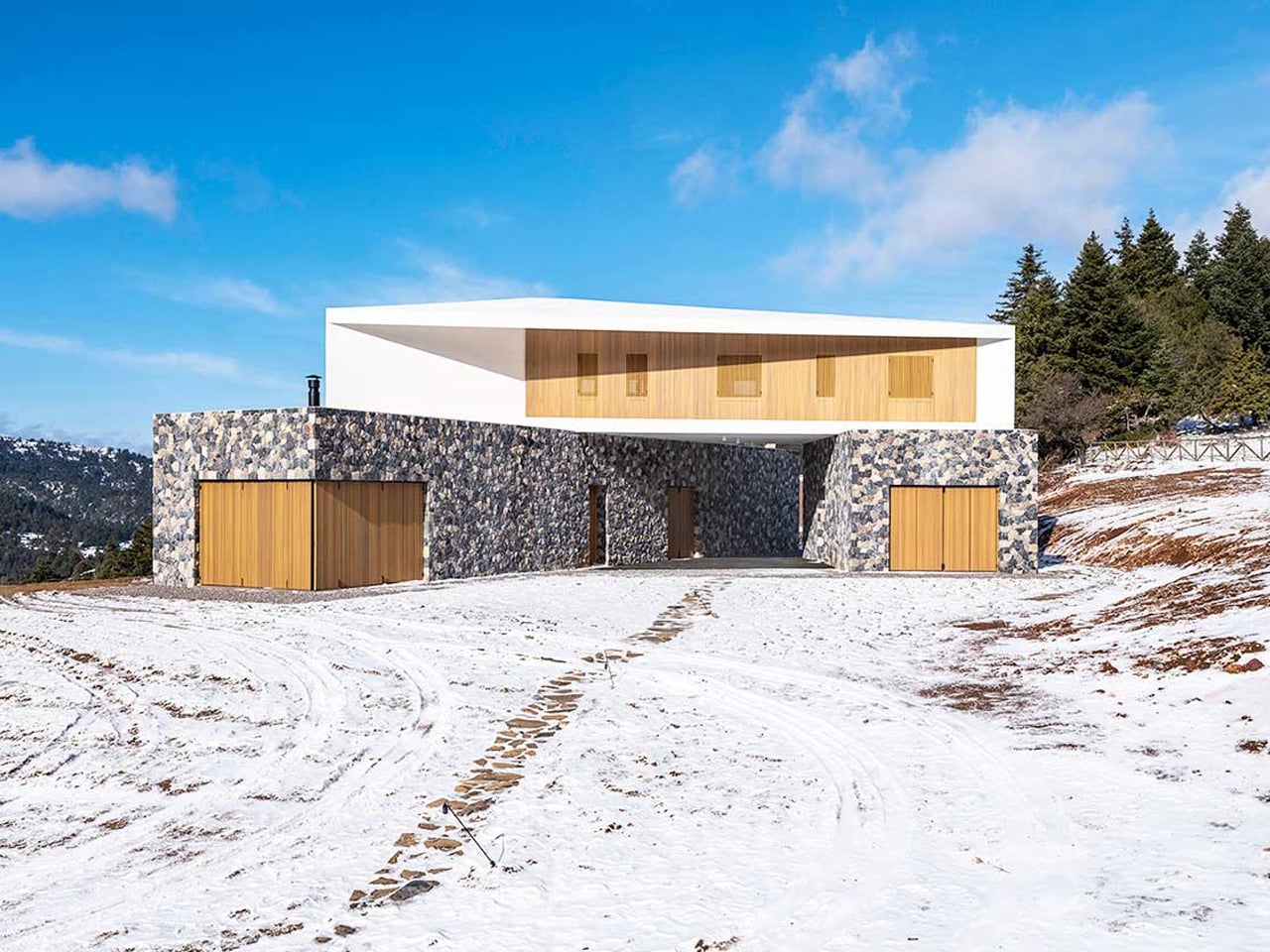 Modern two-story house with a stone base and white upper volume, wooden garage doors, set in a snowy landscape under a blue sky.