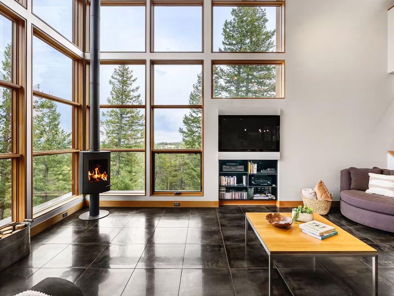 Bright living room with a wall of tall windows, a black wood stove, and a wooden coffee table.