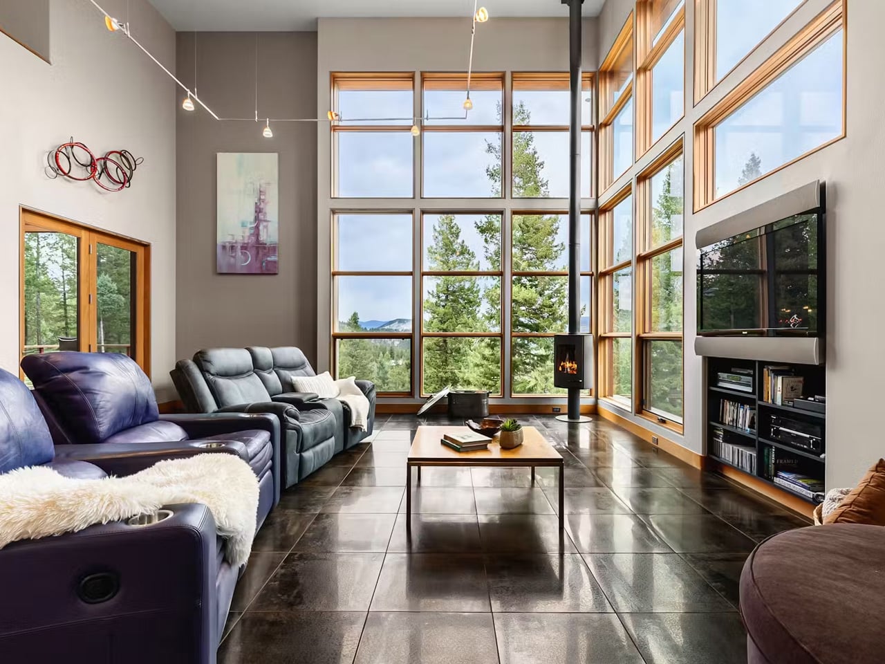 Bright living room with floor-to-ceiling windows overlooking evergreen trees, leather sofas, and a wooden coffee table.