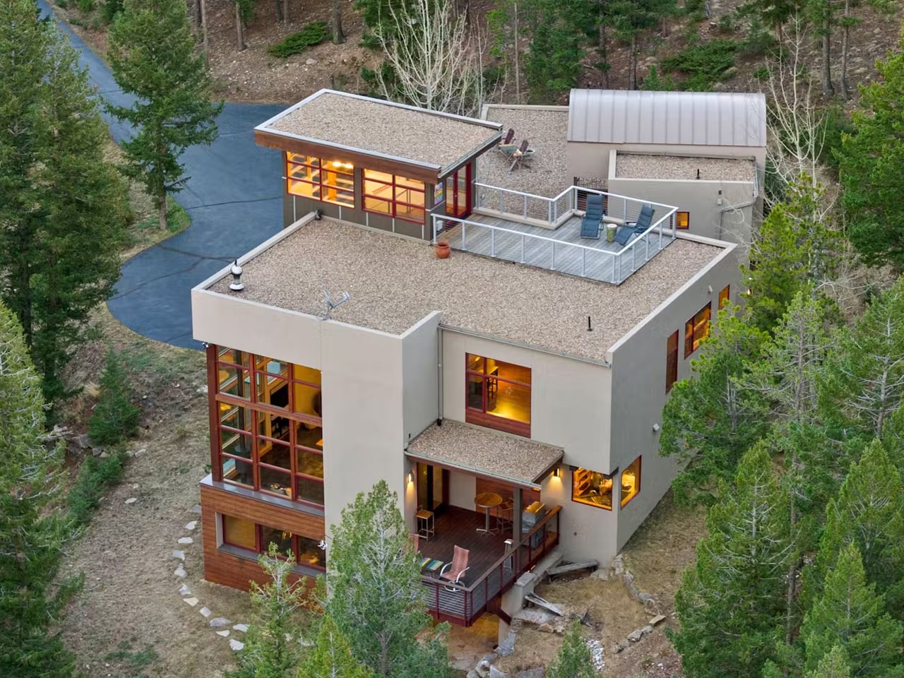 Modern multi-level house with large windows in a pine forest, featuring a rooftop deck and outdoor seating.