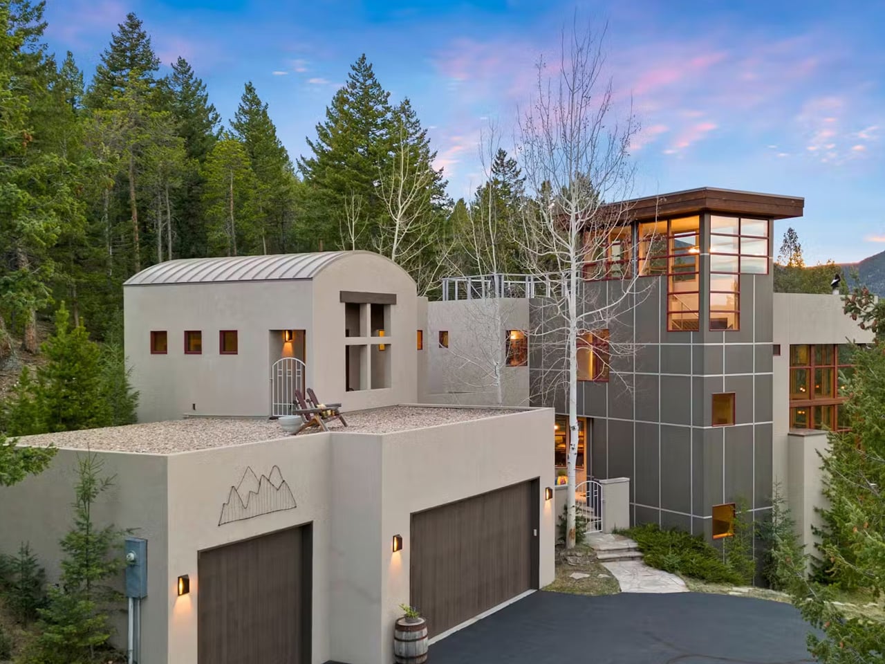 Contemporary multi-story house in a forest, gray exterior, large glass windows, and a rooftop gravel terrace with seating.