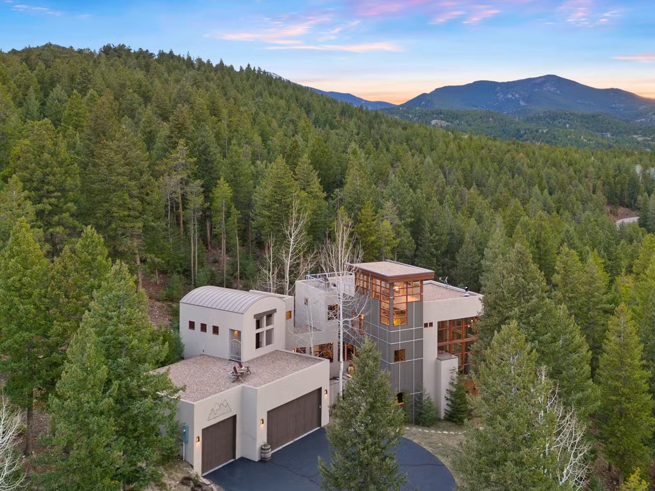 Modern multi‑story house nestled in a dense pine forest with mountains in the background at sunset. A curved-roof garage is visible on the left.