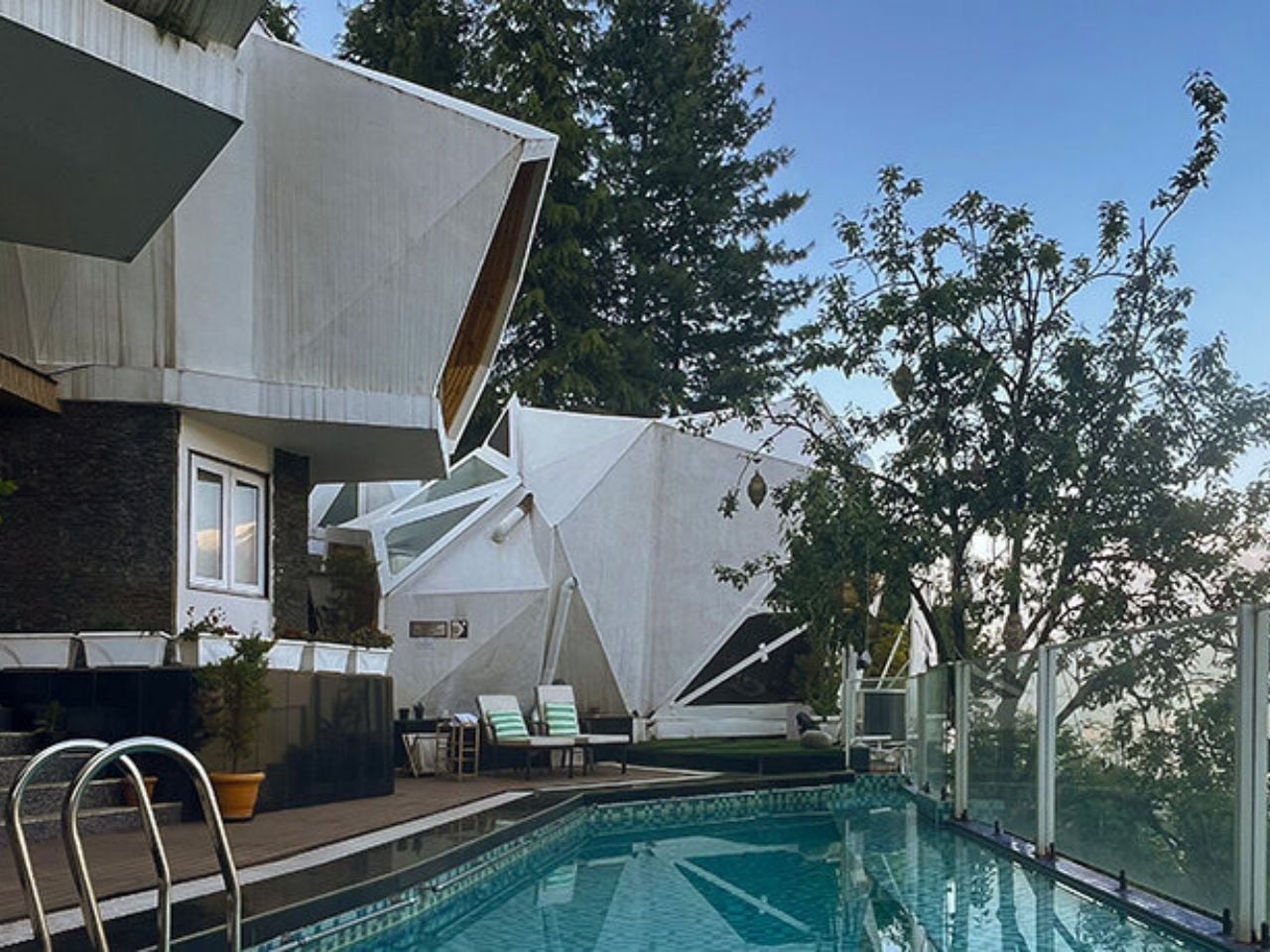 Modern white angular building beside a rectangular pool with glass railing and lounge chairs on a sunny day.