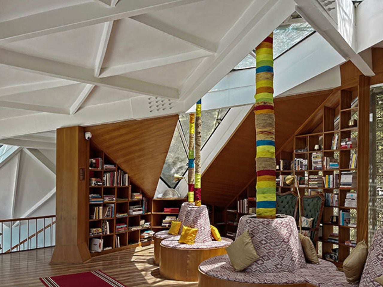 A spacious library lounge with white geometric ceiling, colorful wrapped columns, and circular patterned seating surrounded by bookshelves.