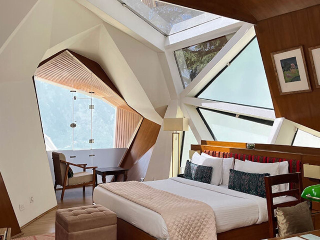 Modern hotel bedroom with a large angular window wall, a king bed with white bedding and decorative pillows, and a seating nook nearby