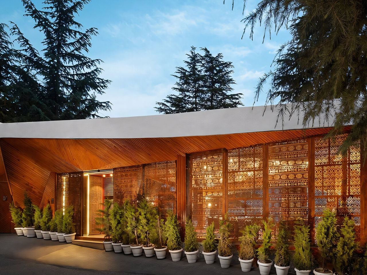 Modern wooden building with glass lattice walls, warm interior lights, and a row of potted evergreens along the entrance.