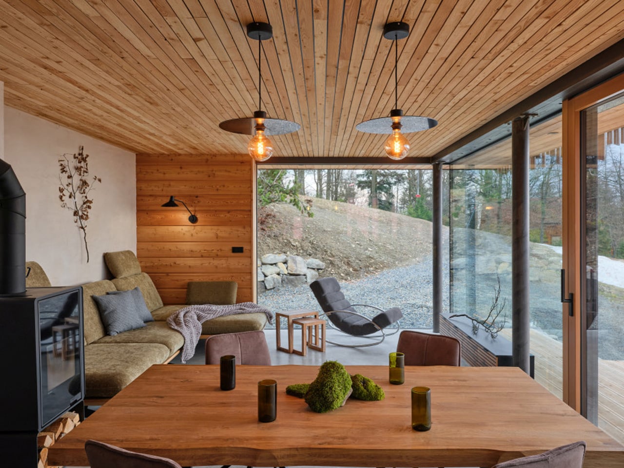 Cozy modern living room with wood panel ceiling and a large glass wall opening to a rocky outdoor area; a wooden dining table with moss centerpiece in foreground and a beige sofa area to the left.