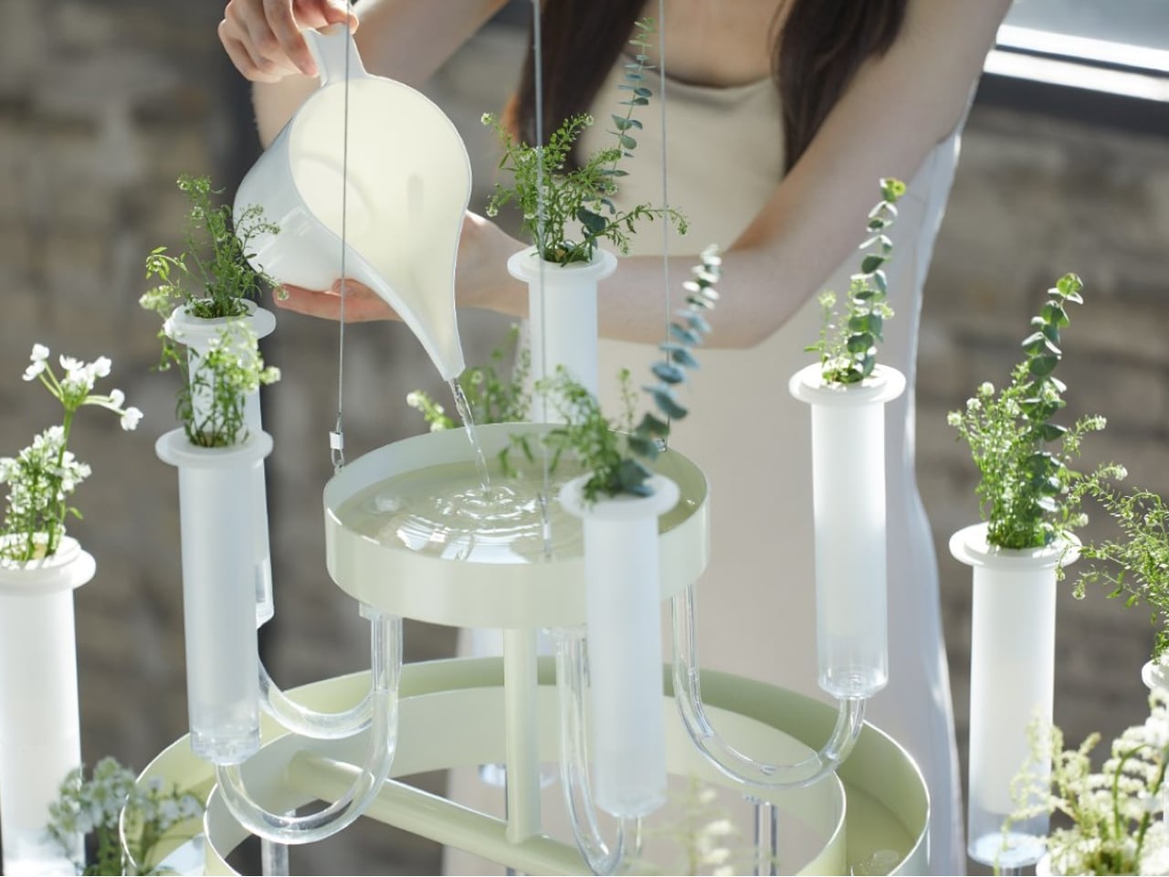 Hands pouring liquid into a pale green chandelier-style planter with glass tubes and small potted plants, an artistic hydroponic display