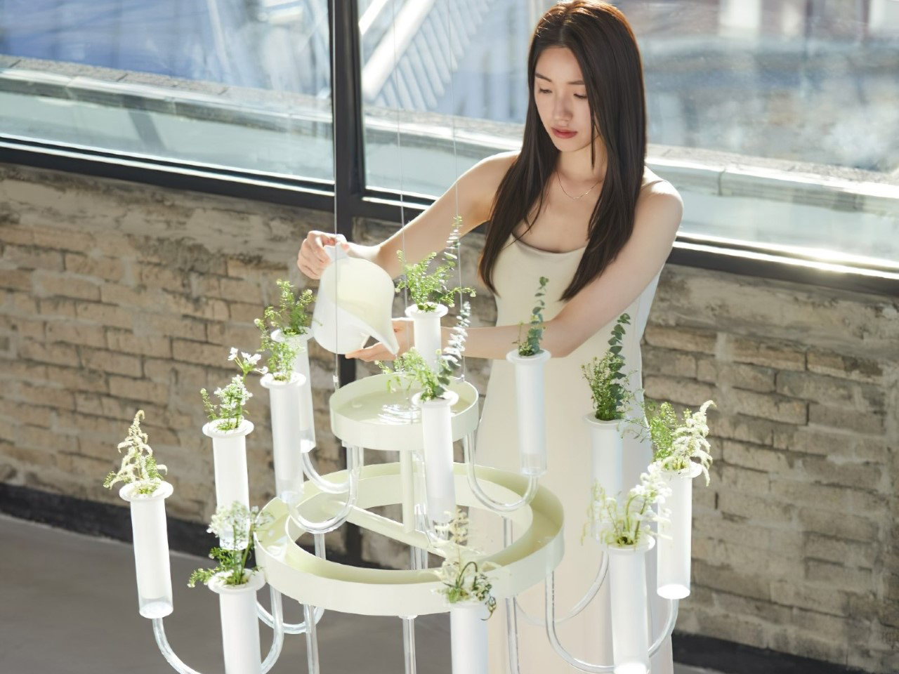 Woman watering a white, plant-filled chandelier indoors by a window.