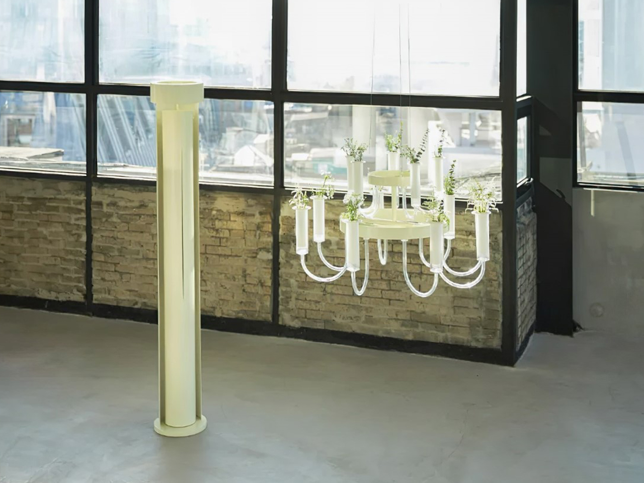 White chandelier with small potted plants on each arm hanging beside a pale yellow floor pillar in an industrial loft with brick wall windows.