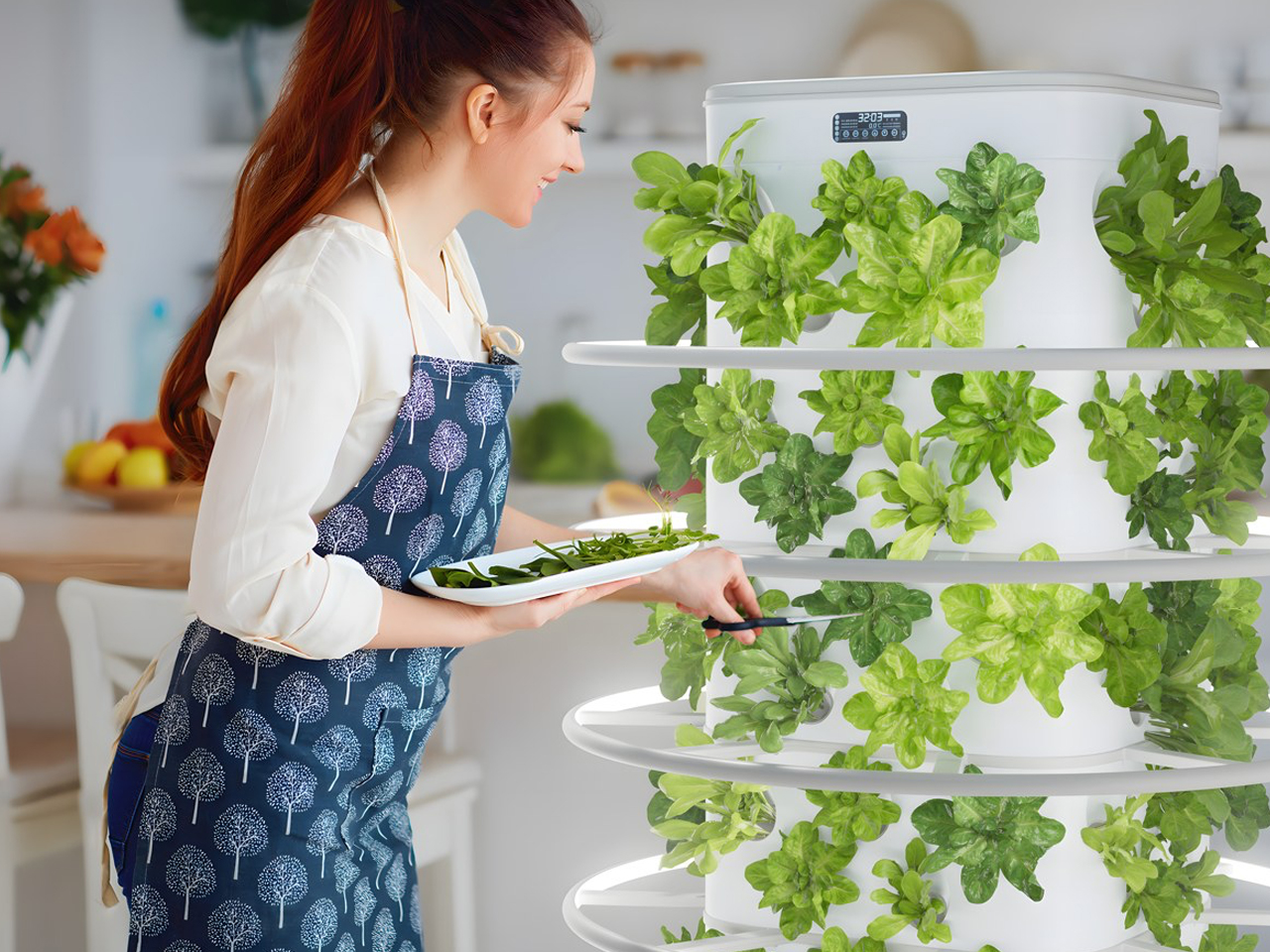 Woman in a blue apron harvests leafy greens from a white multi-tier indoor herb garden, placing herbs on a plate in a bright kitchen.