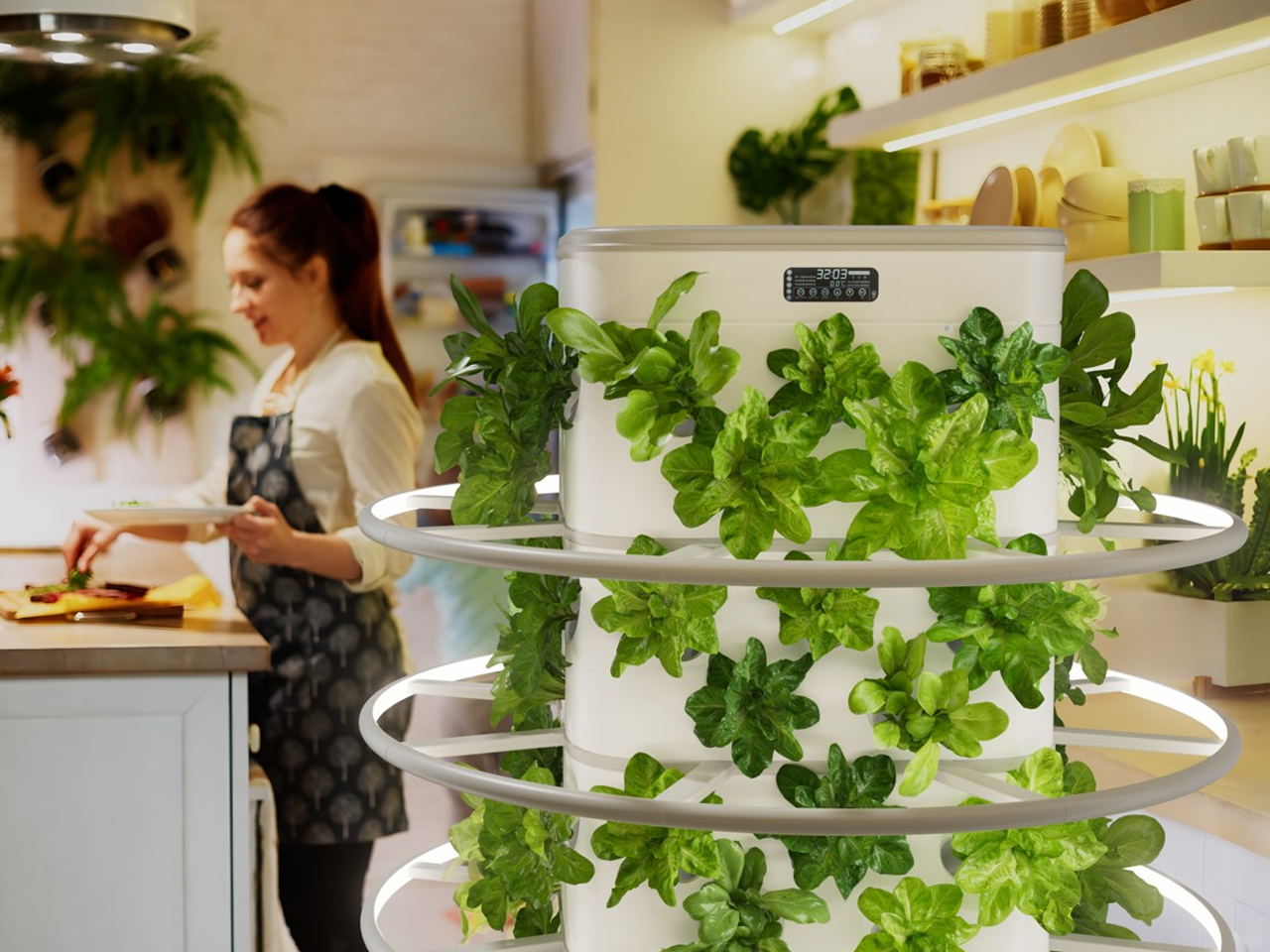 Cook in an apron prepares food at a bright kitchen counter beside a white circular herb garden tower with green leaves.