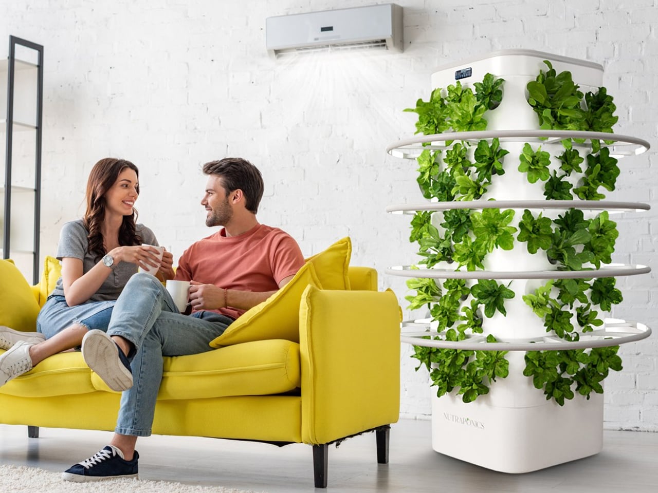 Couple relaxing on a yellow sofa with coffee mugs beside a tall indoor garden tower with leafy greens.