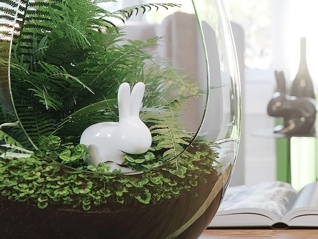 White ceramic bunny nestled among green ferns inside a glass terrarium, on a wooden surface with an open book in the background.
