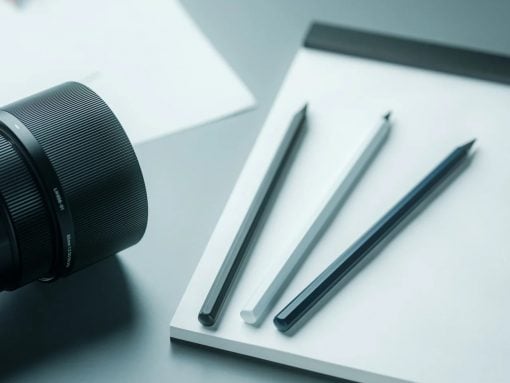Close-up of a camera lens next to a white notebook and three pencils on a light desk surface.