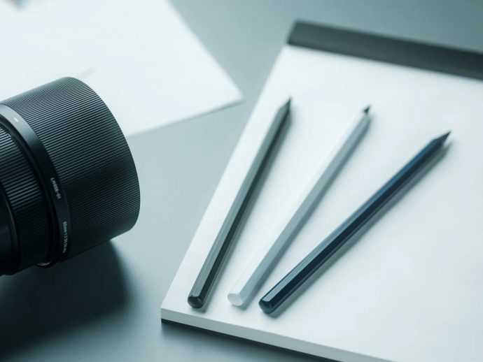 Close-up of a camera lens next to a white notebook and three pencils on a light desk surface.