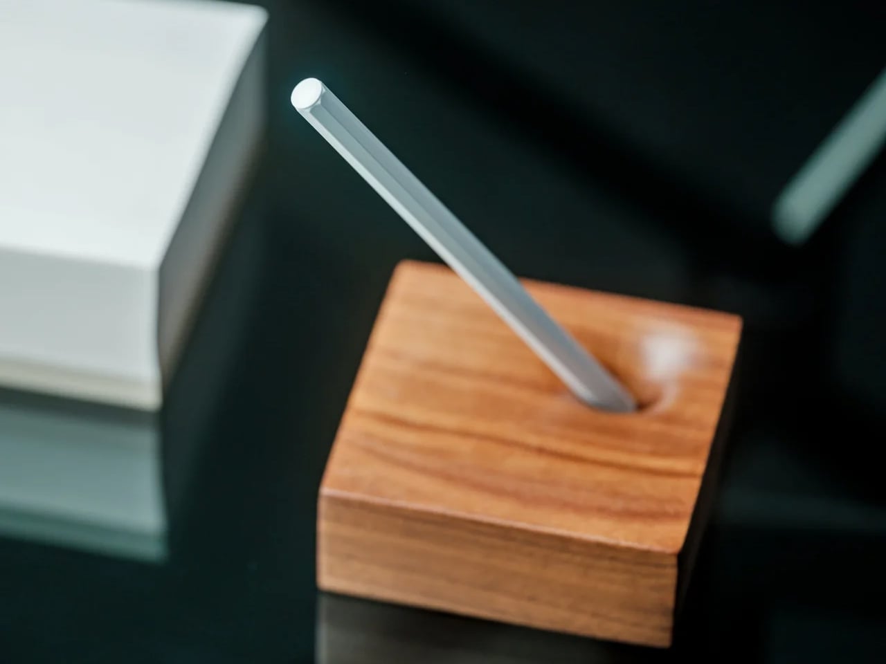 Silver metal rod inserted diagonally through a small square wooden block on a dark surface, like a desk accessory.