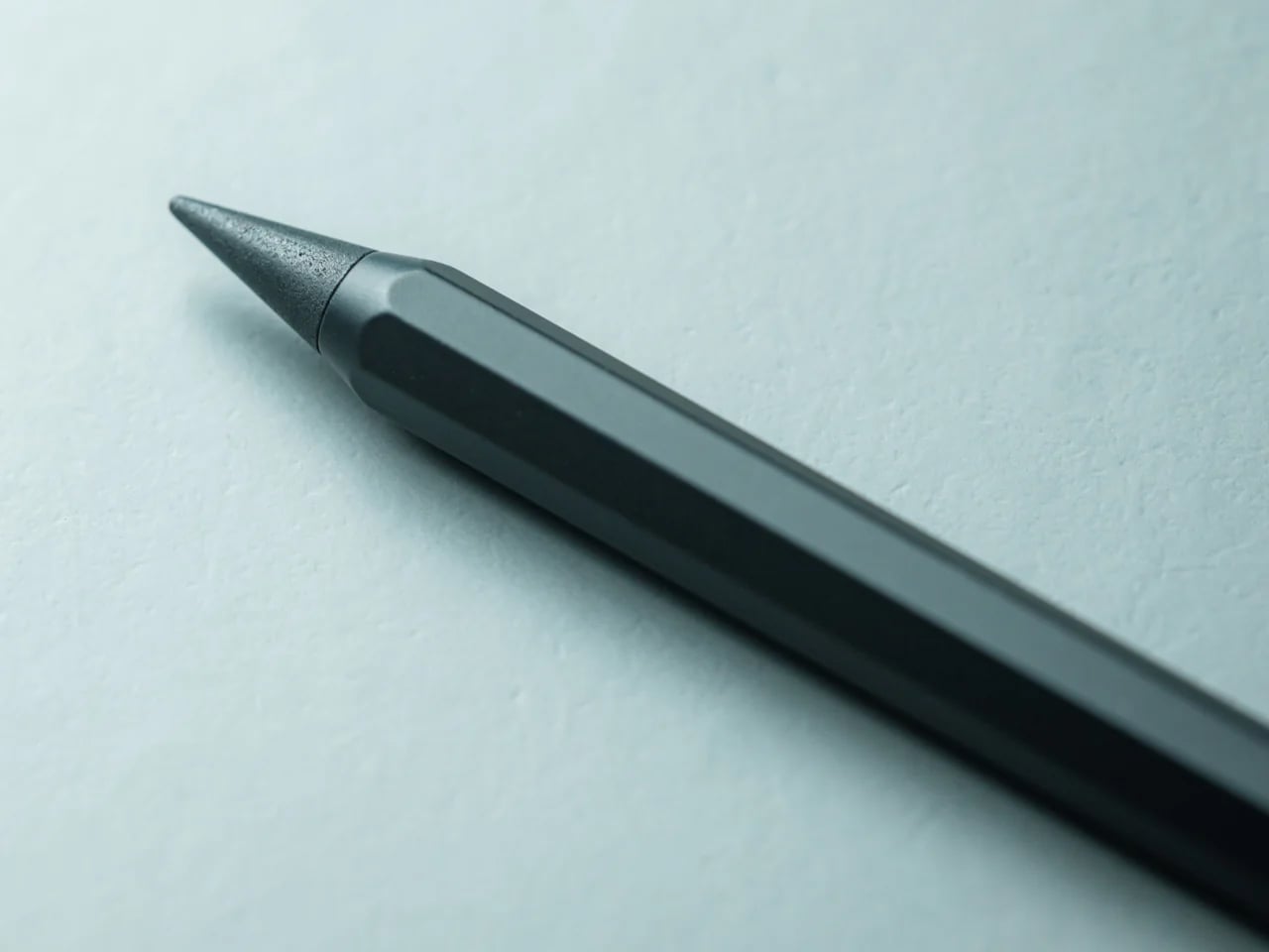 Close-up of a dark pencil-like stylus tip on a light blue background, angled to show the pointed metal nib.