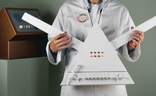 Scientist in a lab coat holds a white triangular electronic instrument with two long grid-like necks and a row of orange buttons, in a studio setting.