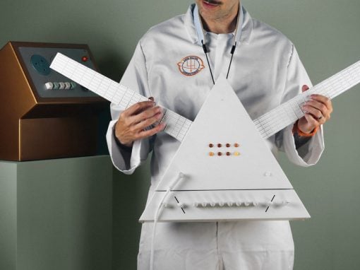 Scientist in a lab coat holds a white triangular electronic instrument with two long grid-like necks and a row of orange buttons, in a studio setting.