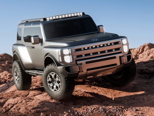 Silver rugged SUV with a roof rack and oversized tires navigating rocky desert terrain under a clear blue sky, front-right angle view.