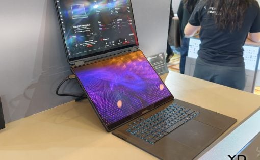 Convertible laptop with a secondary screen upright on a stand at a tech booth, blue-backlit keyboard and display panels visible; person in a dark shirt in the background.