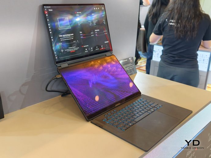 Convertible laptop with a secondary screen upright on a stand at a tech booth, blue-backlit keyboard and display panels visible; person in a dark shirt in the background.