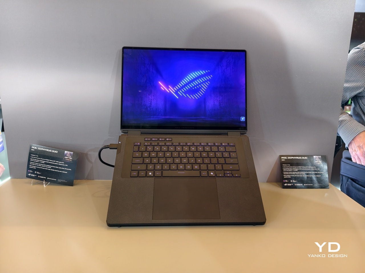 Open dual-screen laptop on a showroom table with purple backlit keyboard and a blue neon screen display on the main panel.
