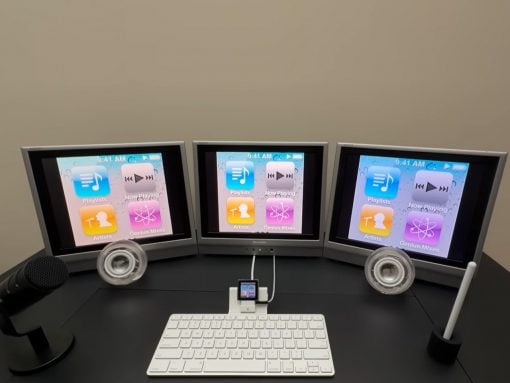 Three-monitor computer desk setup displaying iTunes app icons on all screens, with a microphone on the left and a white keyboard in front.