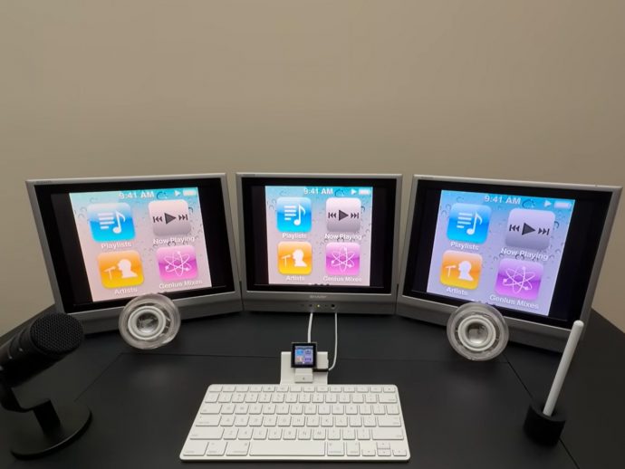 Three-monitor computer desk setup displaying iTunes app icons on all screens, with a microphone on the left and a white keyboard in front.