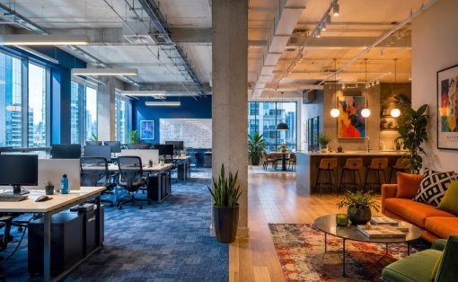 Open-plan office with blue accent walls, row desks on the left, and a cozy lounge with an orange sofa and colorful art on the right.