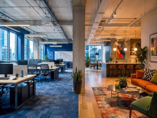 Open-plan office with blue accent walls, row desks on the left, and a cozy lounge with an orange sofa and colorful art on the right.