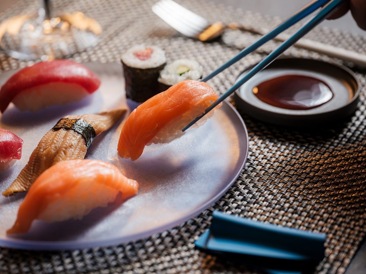 Salmon nigiri being picked up by chopsticks over a plate of assorted sushi and soy sauce nearby.